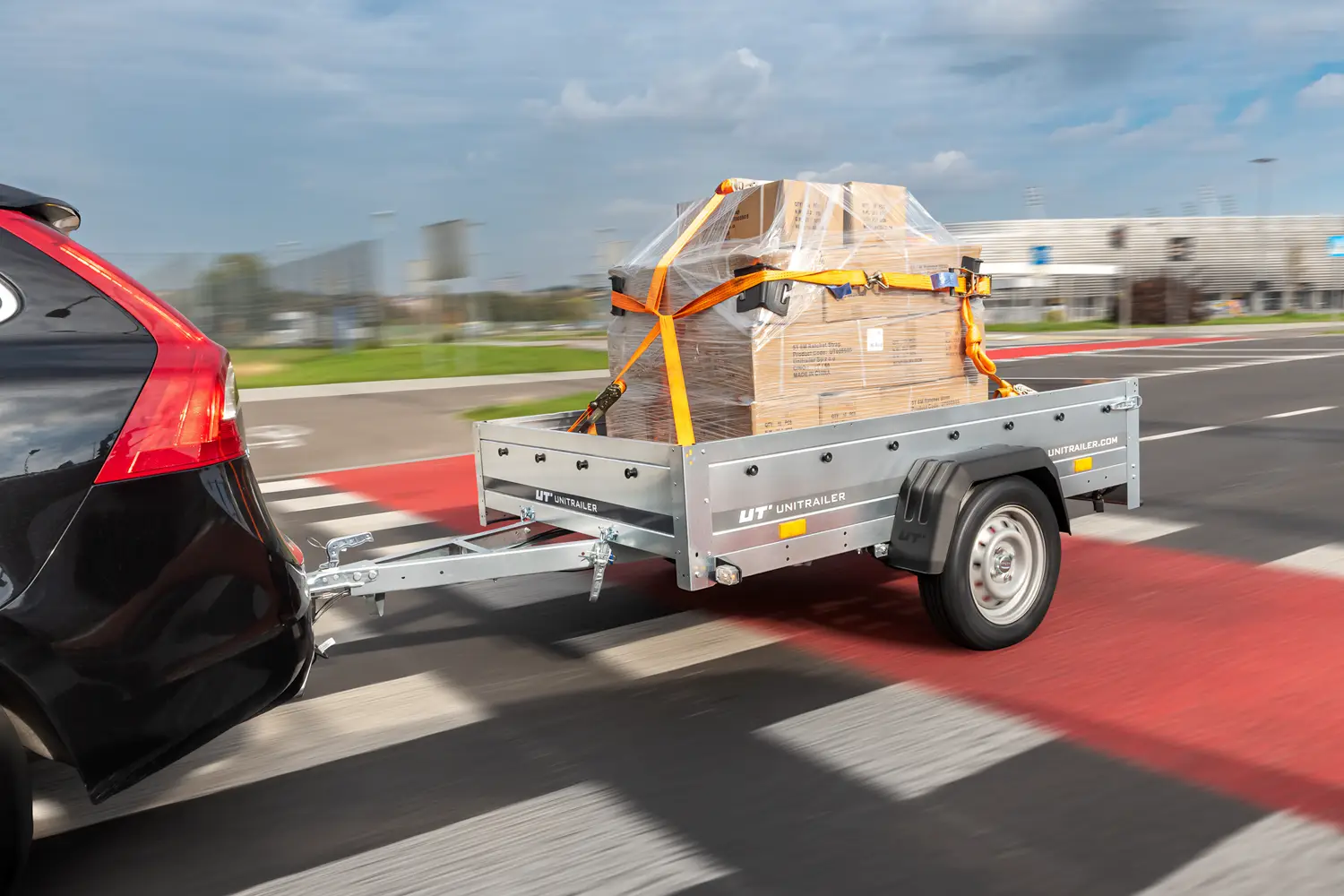 Rimorchio da trasporto Unitrailer agganciato a un'auto nera, che trasporta un bancale con scatoloni avvolto in film estensibile e fissato con cinghie di ancoraggio arancioni durante la marcia su strada