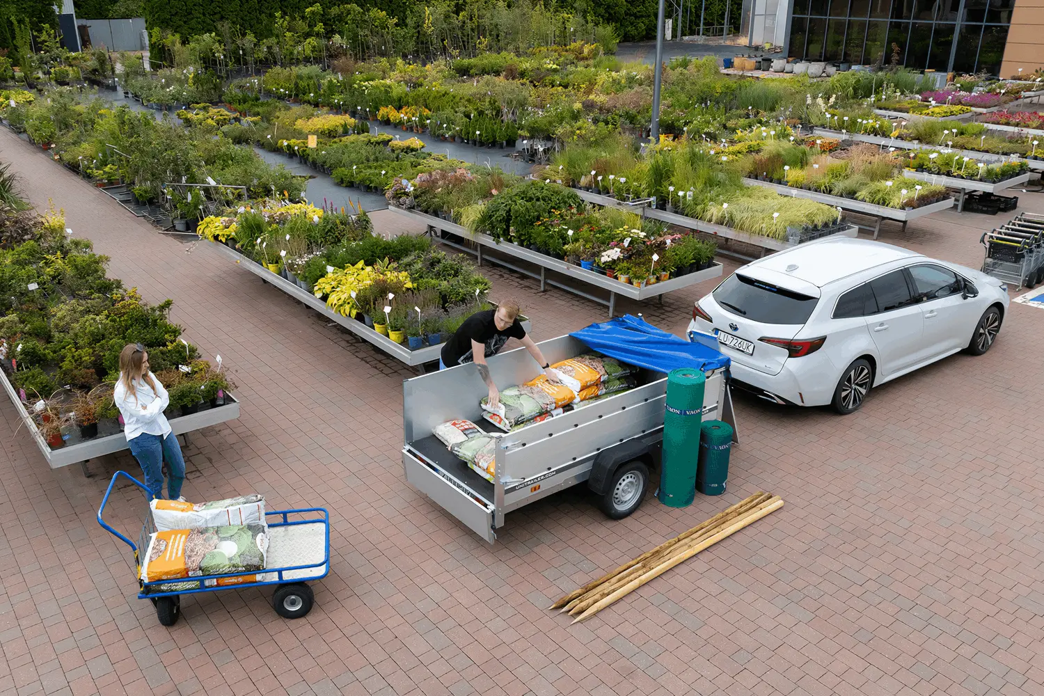 Vista dall'alto di un garden center dove un'auto bianca con rimorchio Unitrailer viene caricata con sacchi di terriccio e materiali per il giardino.